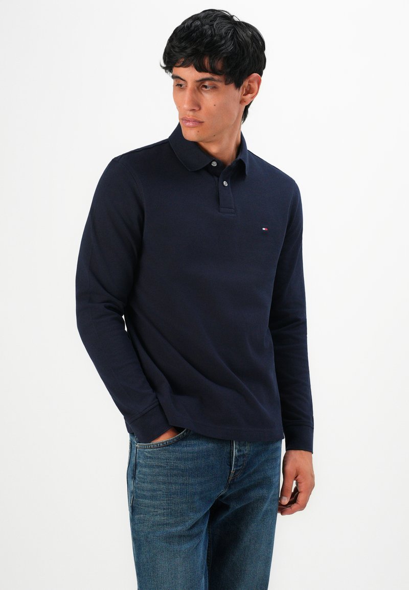 Man i marinblå långärmad polo skjorta och blå jeans, stående med en hand i fickan, tittande åt vänster mot en vit bakgrund.