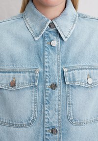 Lichtblauwe spijkerjack met een knoopsluiting aan de voorkant, twee borstzakken, metalen knopen met een logo en een puntige kraag.