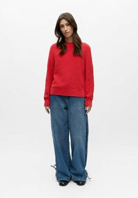 Suéter rojo de punto con mangas largas, combinado con vaqueros de mezclilla azul claro de pierna ancha, con cordones en el dobladillo y aberturas laterales.