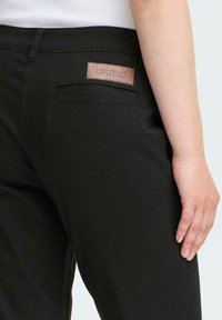 Close-up of a person wearing black pants with a back pocket and a brown leather patch labeled "OXMO" near the waistband.
