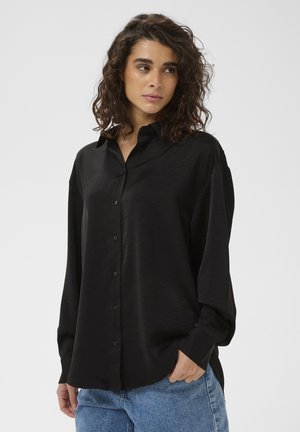 Žena s kučeravými vlasmi v ľahkej čiernej košeli na gombíky a modrých rifliach, stojaca s jednou rukou v vrecku na jednoduchom pozadí.