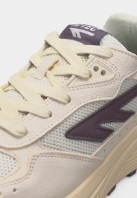 Tenisica bež i svijetlo ljubičaste boje s mrežastim i kožnim panelima, kremastim vezicama i vidljivim logotipom marke na jeziku i bočnoj strani.