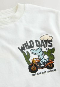 Hvid sweatshirt med et tegneseriealligator på en motorcykel, med kaktuselementer og teksten "VILDE DAGE" og "HAV DIN BEDSTE EVENTYR."