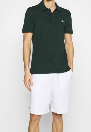 Mand iført mørkegrøn polo og hvide shorts, står med den ene hånd i lommen mod en ensfarvet baggrund.