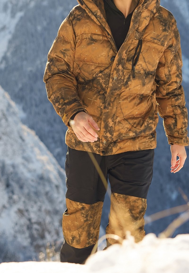 Orange und braune Camouflage-Puffermantel mit Kapuze, ausgestattet mit Reißverschlusstaschen, getragen mit schwarzen Hosen, vor einer verschneiten Bergkulisse.