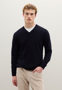 Junger Mann mit kurzem blondem Haar, der einen schwarzen V-Ausschnitt-Pullover über einem weißen Hemd und beigefarbenen Hosen trägt, steht vor einer schlichten hellen Wand.