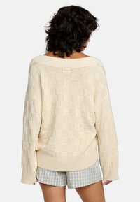 Cremfarbener gestrickter Pullover mit Karomuster, weitem Ausschnitt und gerippten Bündchen. Getragen über karierten Shorts.