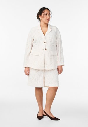 Kvinde iført en hvid blonderblazer og matchende knælange shorts med sorte slingback-hæle, der kigger til sin venstre side.