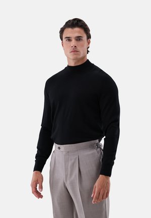 Mann in schwarzem Rollkragenpullover und hochgeschnittenen beigen, plissierten Hosen vor einem einfarbigen Hintergrund.