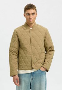 Młody mężczyzna z krótkimi brązowymi włosami, ubrany w pikowaną beżową kurtkę na białą koszulę i jasnoniebieskie dżinsy, z jedną ręką w kieszeni, o neutralnym wyrazie twarzy.