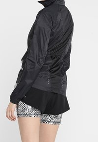 Zwarte lichte jas met hoge kraag, elastische tailleband en mesh achterkant, gecombineerd met zwart-wit gemusterde shorts.