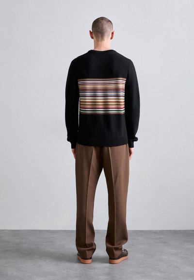 Black knit sweater with a multicolored stripe pattern across the back, paired with loose brown trousers and dark shoes.
