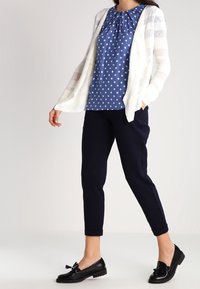 Blå blus med prickar, rynkad halsringning, vit randig kofta, marinblå byxor och svarta slip-on skor med en rosett.