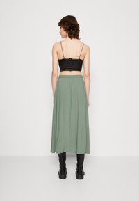Un crop top noir à fines bretelles, associé à une jupe verte fluide arrivant aux chevilles ; le modèle porte des bottes noires hautes.