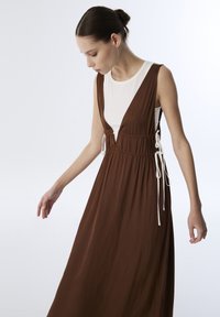 Bruine maxi-jurk met een diepe V-hals en zijvergarningen, met witte strikjes in de taille. De stof lijkt glad en lichtgewicht te zijn.