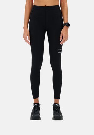 Person wearing black athletic leggings with white logo on left thigh, black running shoes, and a black wristwatch, standing against white background.