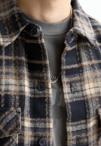 Gros plan sur une personne portant une chemise à carreaux beige et marine, boutonnée, sur un t-shirt gris à col rond, avec une chaîne en argent.