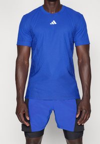 Mann trägt ein blaues Sportshirt und passende Shorts mit schwarzen Einsätzen und Adidas-Logo auf der Brust vor weißem Hintergrund.