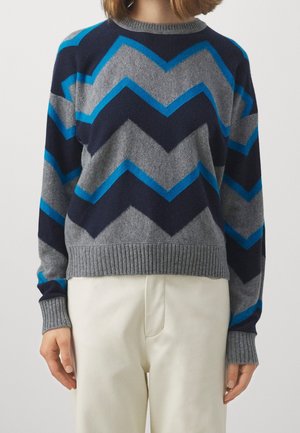 Personne portant un pull à motif chevron gris, marine et bleu avec poignets et ourlet côtelés, associé à un pantalon clair.