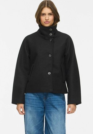 Femme portant une veste noire à boutons avec un col montant et un jean bleu, debout devant un fond clair uni.