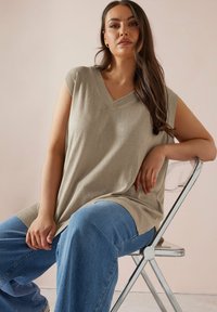 Beige stickad tunika med V-hals och korta ärmar, kombinerad med blå vida jeans. Modellen sitter på en genomskinlig stol mot en neutral bakgrund.