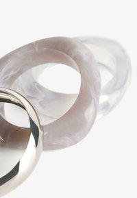 Három gyűrű: egy fényes fémből készült szalag, egy áttetsző, világos rózsaszínű, örvénylő mintával, és egy tiszta, fényes darab. Simára csiszolt felületek és lekerekített formák.