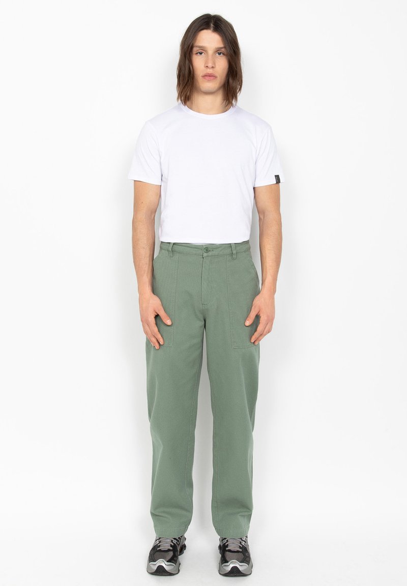 Jeune homme debout, portant un t-shirt blanc uni, un pantalon vert ample avec de grandes poches, et des baskets noir-gris sur un fond blanc.