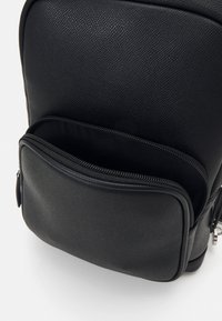 Svart ryggsekk laget av teksturert kunstskinn, med en frontlomme med glidelås og glatte kanter. Elegant, minimalistisk design.