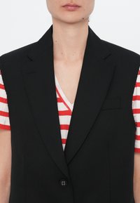 Juodas pritaikytas liemeniškas švarkas su ryškiomis atlapėmis, užsegamas vienu mygtuku, su lygia tekstūra, dėvimas ant raudono ir baltos juostelėmis marškinėlių.