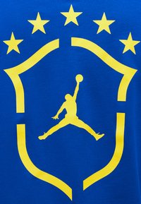 Κίτρινη σιλουέτα παίκτη μπάσκετ που πηδάει με την μπάλα μέσα σε μια ασπίδα, με πέντε κίτρινα αστέρια στην κορυφή, σε μπλε φόντο.