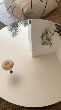 Mesa de café redonda branca com uma superfície lisa, uma peça de destaque em bambu tecido e uma pequena planta refletida no acabamento brilhante.