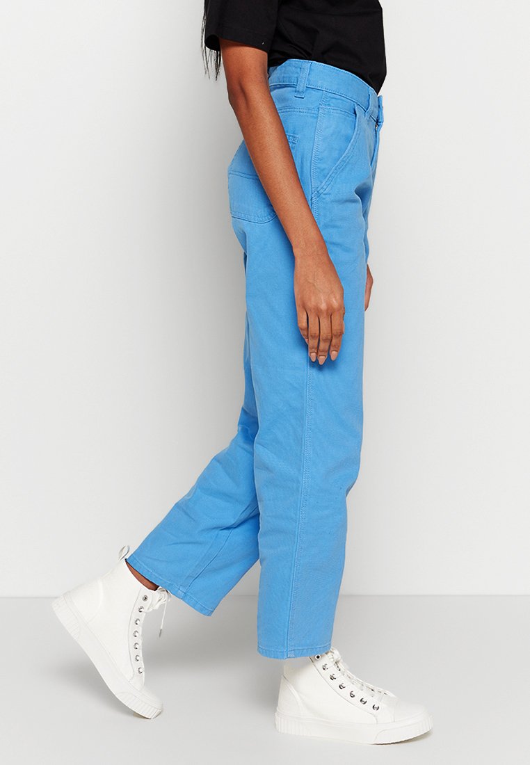 Pantaloni di cotone blu chiaro con taglio dritto, tasche laterali e passanti per cintura, abbinati a sneaker alte bianche.