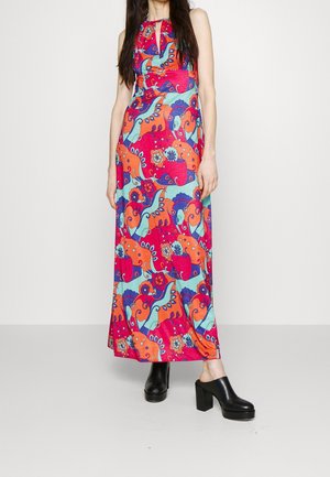 Vrouw draagt een mouwloze, tot op de grond vallende jurk met een levendig veelkleurig bloemen- en abstract patroon, gecombineerd met zwarte plateau mule schoenen.