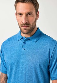 Mies, jolla on lyhyet hiukset ja parta, pukeutuu siniseen kauluspaitapoloon, jossa on hienovarainen brodeerattu logo ja näkyvä käsivarren tatuointi.