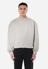 Lys grå oversized sweatshirt med ribbet halsudskæring og lange ærmer, der har subtil branding på brystet, parret med sorte bukser.