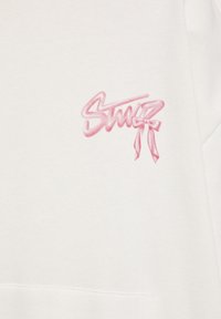 Hvid sweatshirt med pink broderet tekst "Stutz" og en sløjfe detalje. Blød tekstur, afslappet design og minimalistisk stil.