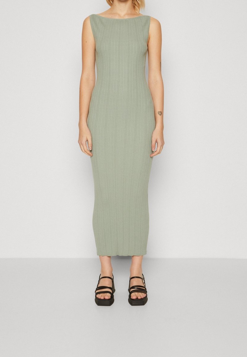 Femme debout vêtue d'une robe midi sans manches côtelée vert clair et de sandales noires à plateforme avec lanières sur un fond uni.