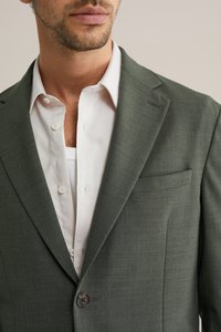 Bărbat purtând un sacou verde închis peste o cămașă albă cu nasturi, cu un maiou alb vizibil la nivelul pieptului.