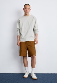 Cămășă lungă gri deschis cu un mic logo Nike, purtată cu pantaloni scurți cargo maro, șosete albe și adidași bej cu talpă din cauciuc.