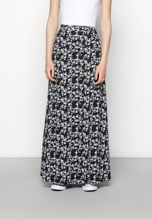 Woman wearing a black maxi skirt with white floral patterns, white sleeveless top, and white sneakers, standing against a plain background.