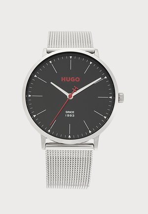 Montre-bracelet à bracelet en maille argentée avec cadran rond noir, aiguilles blanches des heures et des minutes, aiguille des secondes rouge, et logo rouge "HUGO".