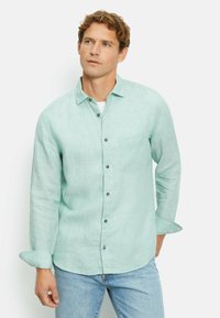 Lichtgroen linnen overhemd met lange mouwen, knoopsluiting aan de voorkant, één borstzak en grijze knopen. Draagt met blauwe jeans.