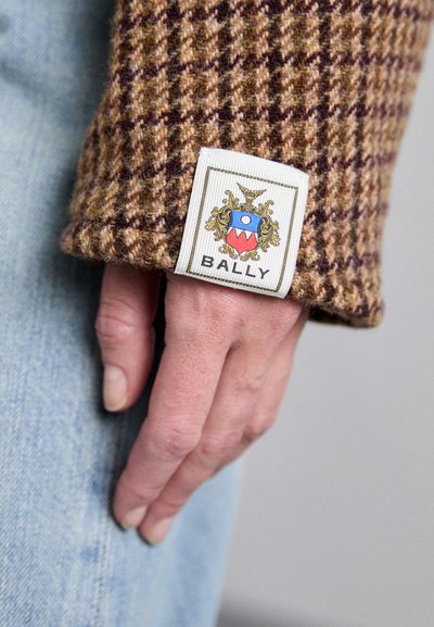 Brun houndstoothærme lavet af tekstureret stof, der har et hvidt mærkat med et farverigt Bally-logo og udsmykkede detaljer.