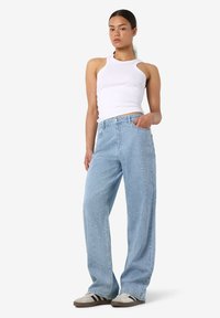 Moteris dėvi baltą asimetrinį berankovį viršų, šviesiai mėlynas laisvas džinsas su mažais papuošimais ir baltus sportbačius su juodomis juostelėmis.