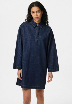 Kvinna med lockigt mörkt hår som bär en lös mörk jeansklänning med lång ärm, krage och knappstängning fram.