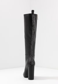 Wysoki czarny skórzany kozak na wysokim obcasie blokowym, z białym zygzakowatym przeszyciem biegnącym pionowo po tylnej stronie.