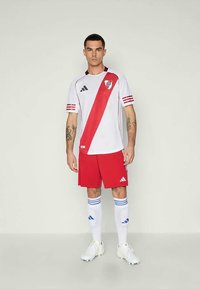 Vit fotbollströja med ett diagonalt rött stripe, röda shorts, vita strumpor med blå ränder och vita fotbollsskor. Adidas-logotyp närvarande.