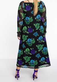Robe noire avec des manches longues transparentes, ornée d'un motif floral de roses bleues, vertes et violettes, s'étendant jusqu'à la cheville. Talons à lanières violets.
