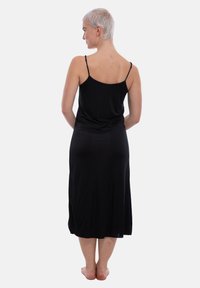 Una donna con capelli biondi corti sta scalza, voltata di spalle, indossando un abito midi nero senza maniche con spalline sottili su uno sfondo semplice.