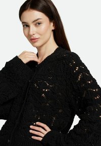 Cardigan noir en maille avec des motifs ajourés complexes, des manches longues, un col rond et une fermeture à boutons à l'avant.